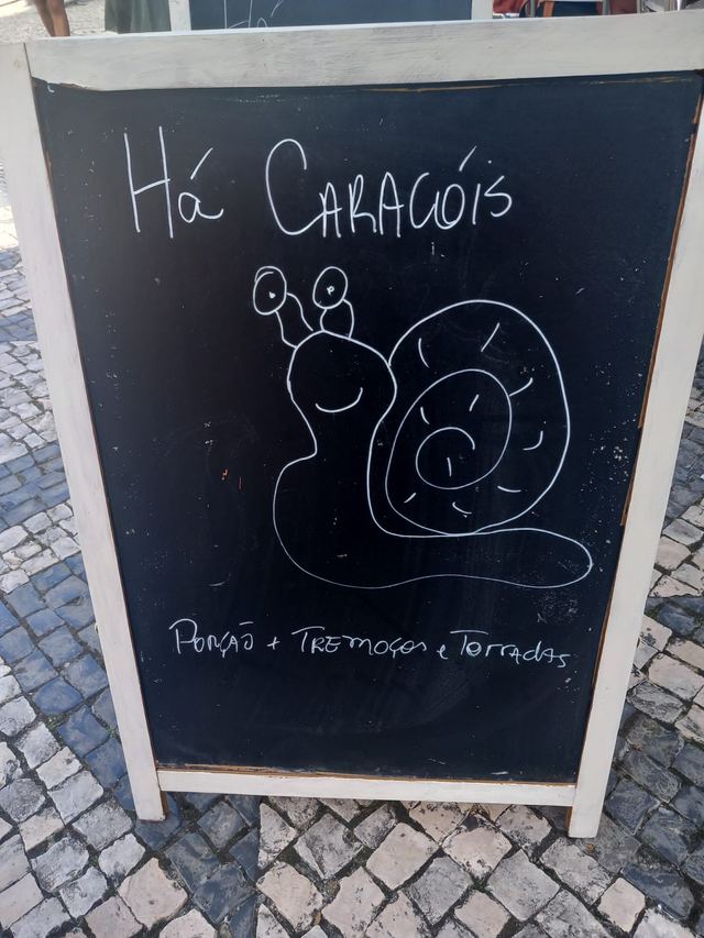 Chalkboard A-frame sign reading “Há Caracóis” above a snail drawn in white chalk, on a cobbled pavement