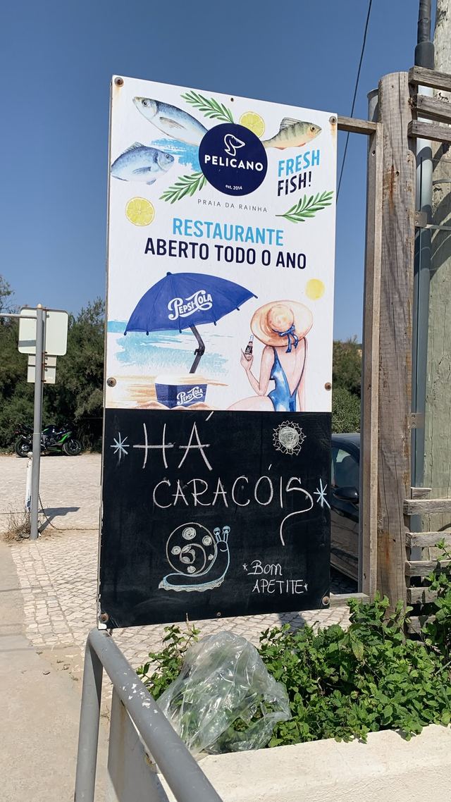 Chalkboard panel under the Pelicano restaurant sign at Praia da Rainha, reading “Há Caracóis” beside a chalk snail and the words “Bom apetite”