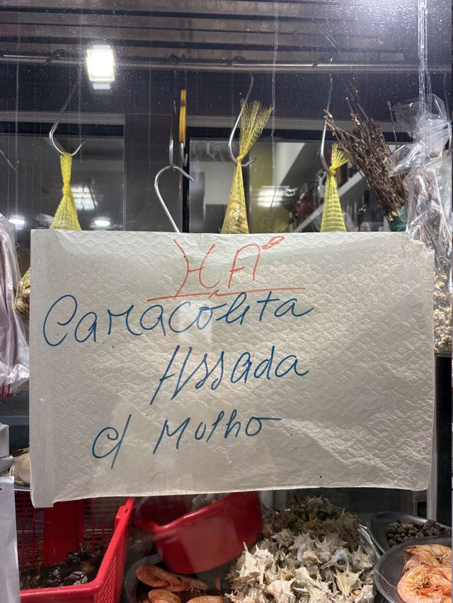 Paper napkin taped to a seafood counter, reading “Há Caracoleta Assada c/ Molho” in red and blue marker