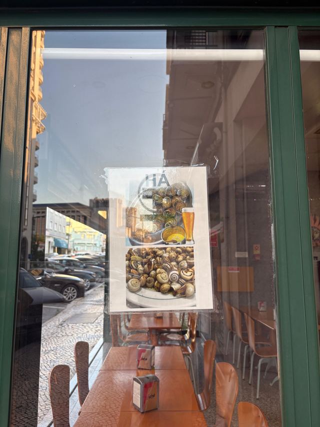 Laminated “Há” poster showing snails and a glass of beer, taped inside the green window frame at Estrela de Vilhena